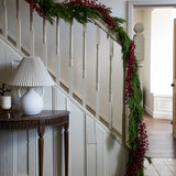 Decorative Christmas garland with red berries on a staircase wall, with a side table and lamp in the foreground.