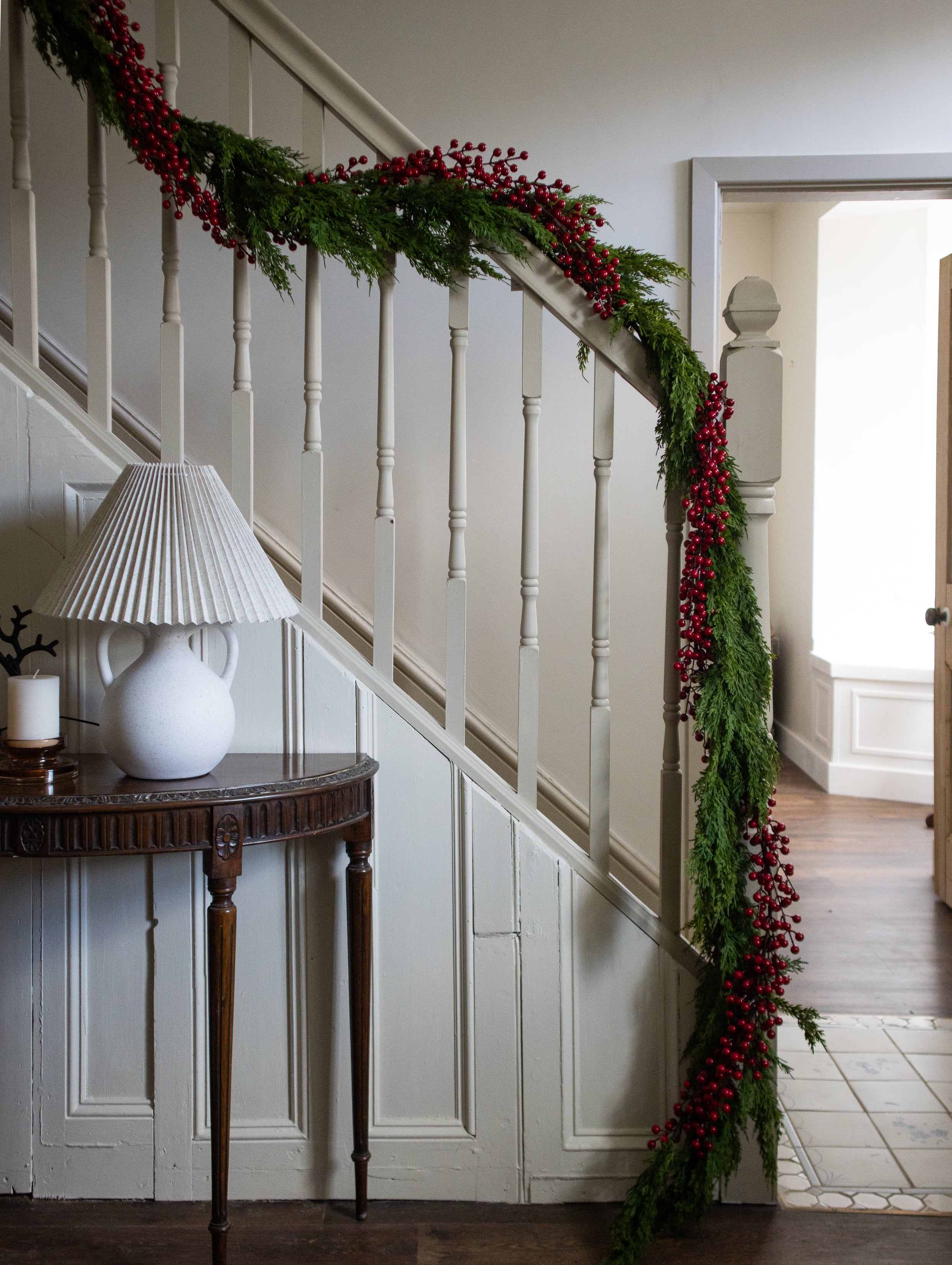 Decorative Christmas garland with red berries on a staircase wall, with a side table and lamp in the foreground.