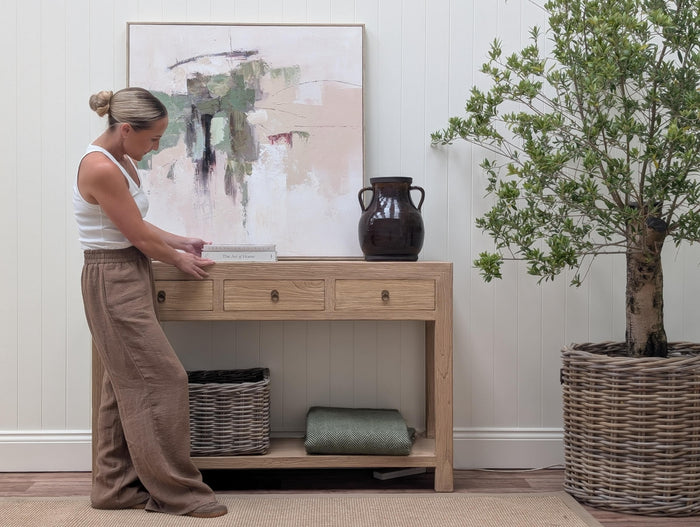 A woman in a white tank top and brown pants stands beside a light wood console table in a styled room. The table has three drawers, an open lower shelf, and is decorated with a dark ceramic vase, a framed abstract print, a wicker basket, and a folded green throw blanket.