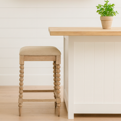 A farmhouse-style kitchen with white shiplap walls, a wooden floating shelf holding a white ceramic pitcher, stacked plates, bowls, and a small potted herb plant. Below, a light wood and white kitchen island with a beige-upholstered stool featuring turned wooden legs sits on a light hardwood floor, accompanied by another potted herb on the island countertop.