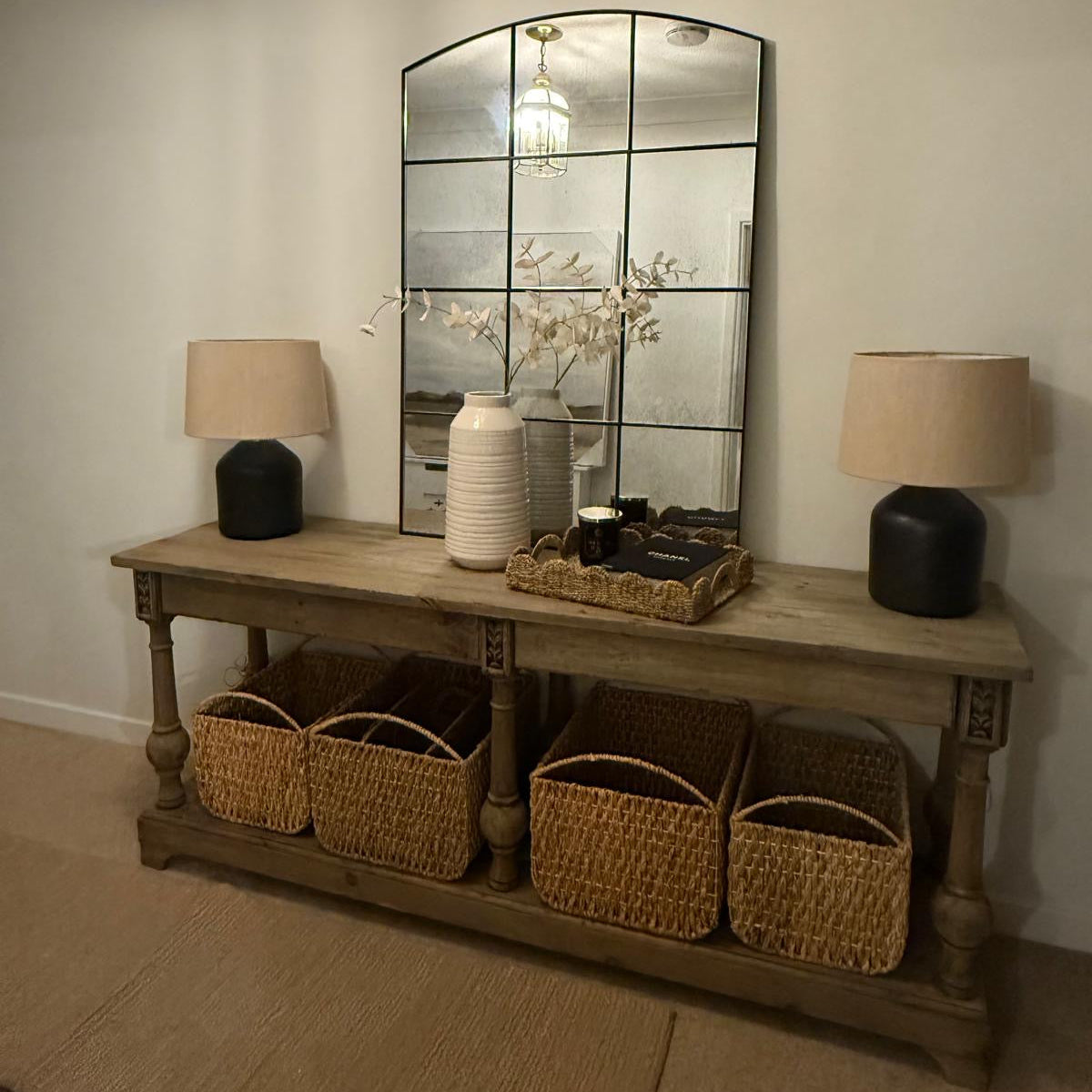 Wooden console table with wicker baskets, lamps, and a decorative mirror in a room.