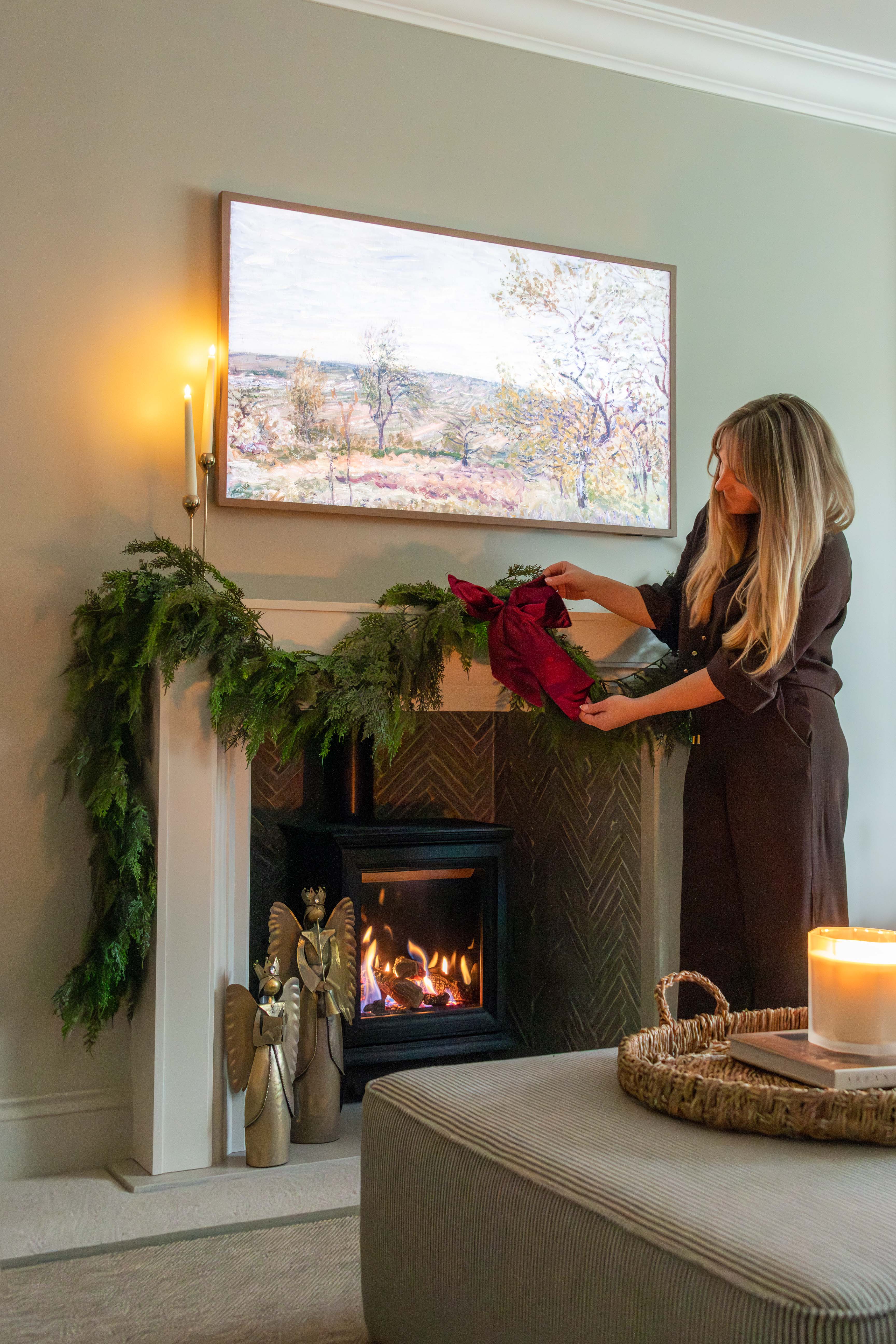 Woman decorating a fireplace with greenery and a red bow in a cozy living room.