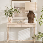 A large lamp with a natural rattan base and a beige linen shade, placed on a white console table in a room with a plant and a framed picture.