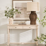 A large lamp with a natural rattan base and a beige linen shade, placed on a white console table in a room with a plant and a framed picture.