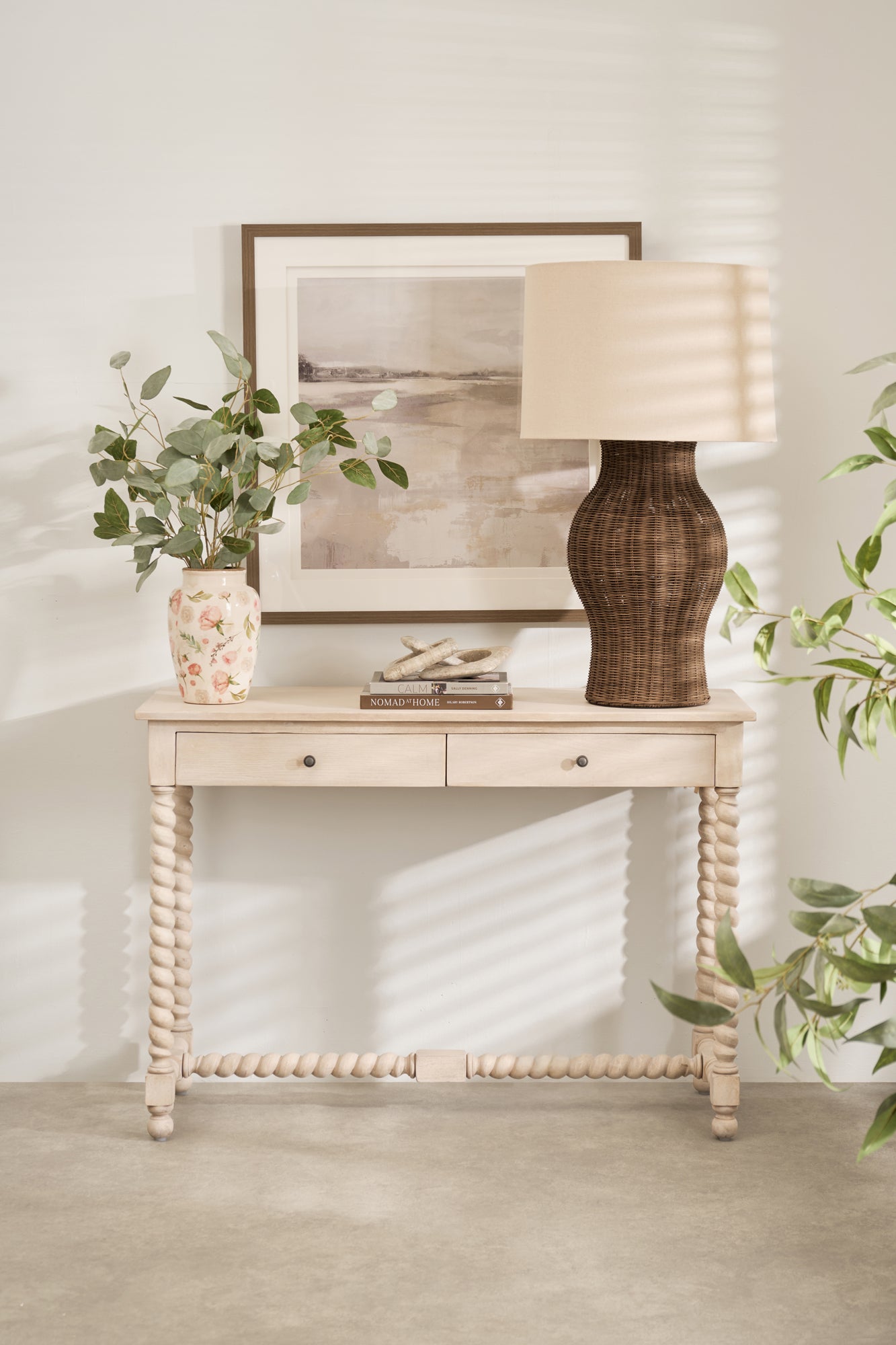 A large lamp with a natural rattan base and a beige linen shade, placed on a white console table in a room with a plant and a framed picture.