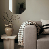 Grey sofa with a striped blanket and pillow, next to a small table with a vase and branches.