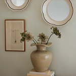 Beige vase with dried flowers on a surface, framed picture, and round mirror on wall.
