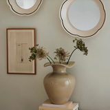 Beige vase with dried flowers on a surface, framed picture, and round mirror on wall.