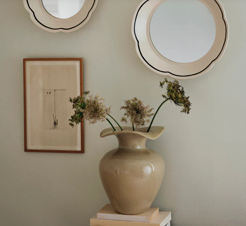 Beige vase with dried flowers on a surface, framed picture, and round mirror on wall.