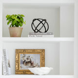 Black metal ring orb decor sculpture on a white shelf beside books and a plant