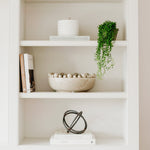 Black metal ring orb decor sculpture on a white shelf beside books and a plant