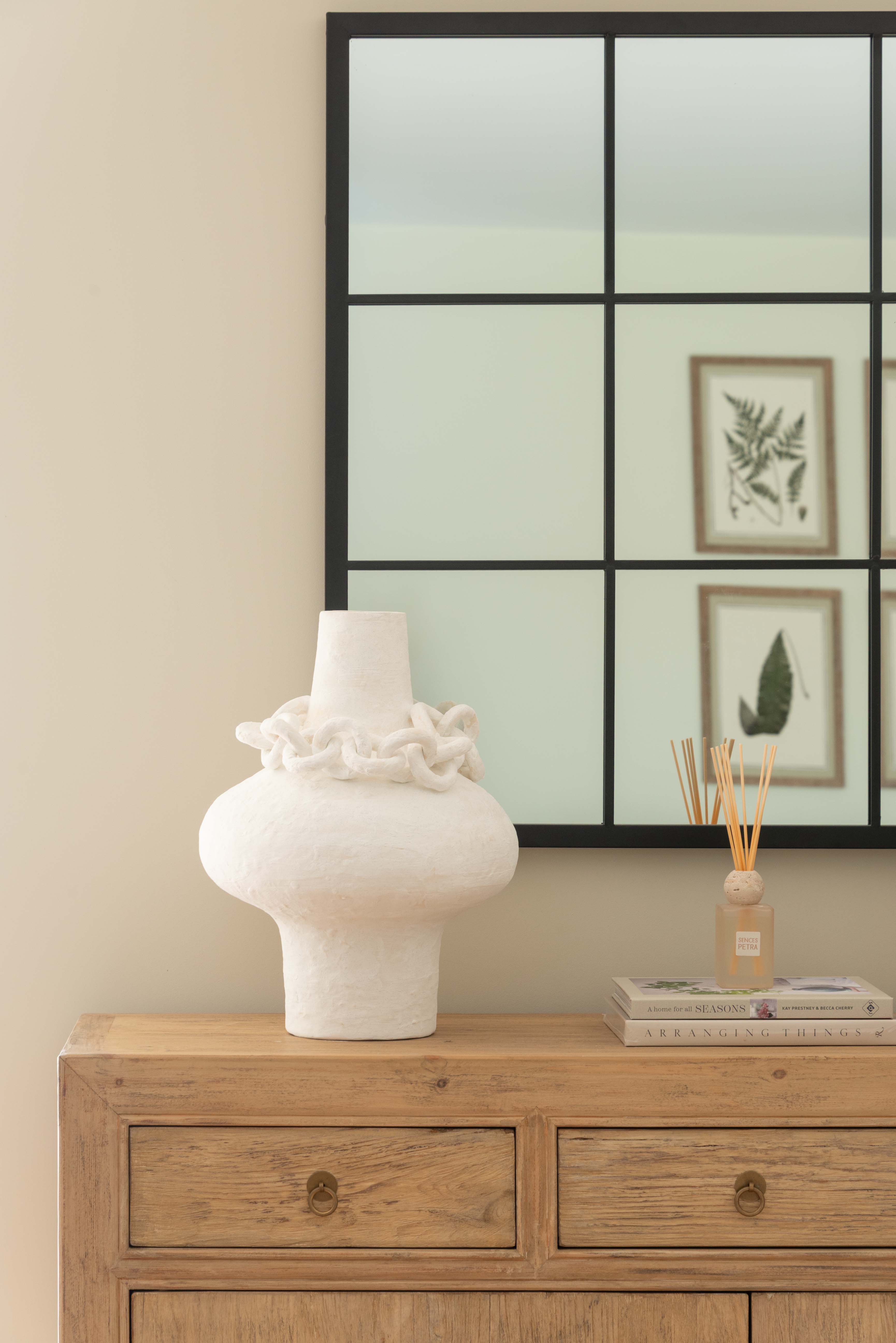 White vase on a wooden dresser with a decorative mirror in the background
