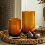 Two orange glass candle holders on a woven tray with figs, against a neutral wall.
