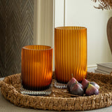 Two orange glass candle holders on a woven tray with figs, against a neutral wall.
