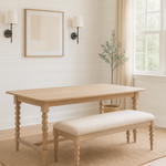A bright dining room with white shiplap walls, a light wood dining table with spindle legs, and a matching beige upholstered bench. The set sits on a textured jute rug over light wood floors. Above the table hangs abstract art flanked by two black sconces with white shades. A large window with beige linen curtains lets in natural light, and an olive tree in a woven basket stands in the corner.