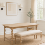 A bright dining room with white shiplap walls, a light wood dining table with spindle legs, and a matching beige upholstered bench. The set sits on a textured jute rug over light wood floors. Above the table hangs abstract art flanked by two black sconces with white shades. A large window with beige linen curtains lets in natural light, and an olive tree in a woven basket stands in the corner.