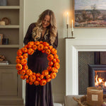 Woman holding a large orange pumpkin wreath in a cozy living room.