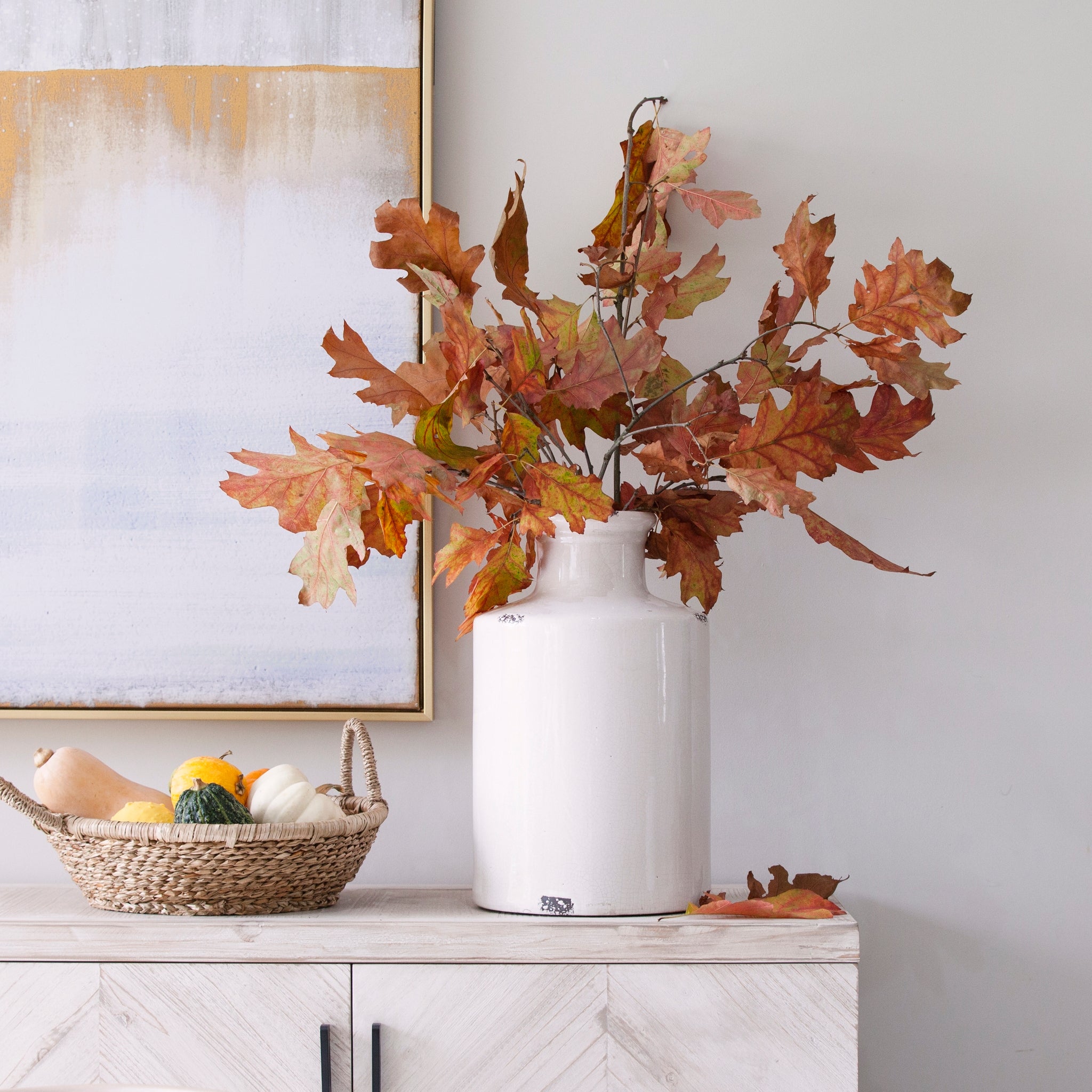White ceramic bottle vase with autumn leaves styled beside a basket of gourds