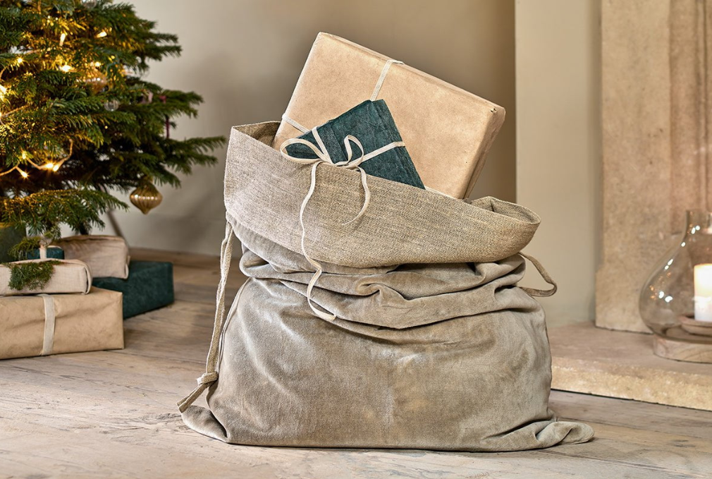 Charles Ted Cotton Velvet Christmas Sack in light grey with gifts inside, styled by a Christmas tree and fireplace