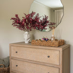 Beige dresser with wicker drawers, decorative flowers, and a mirror on a beige wall.