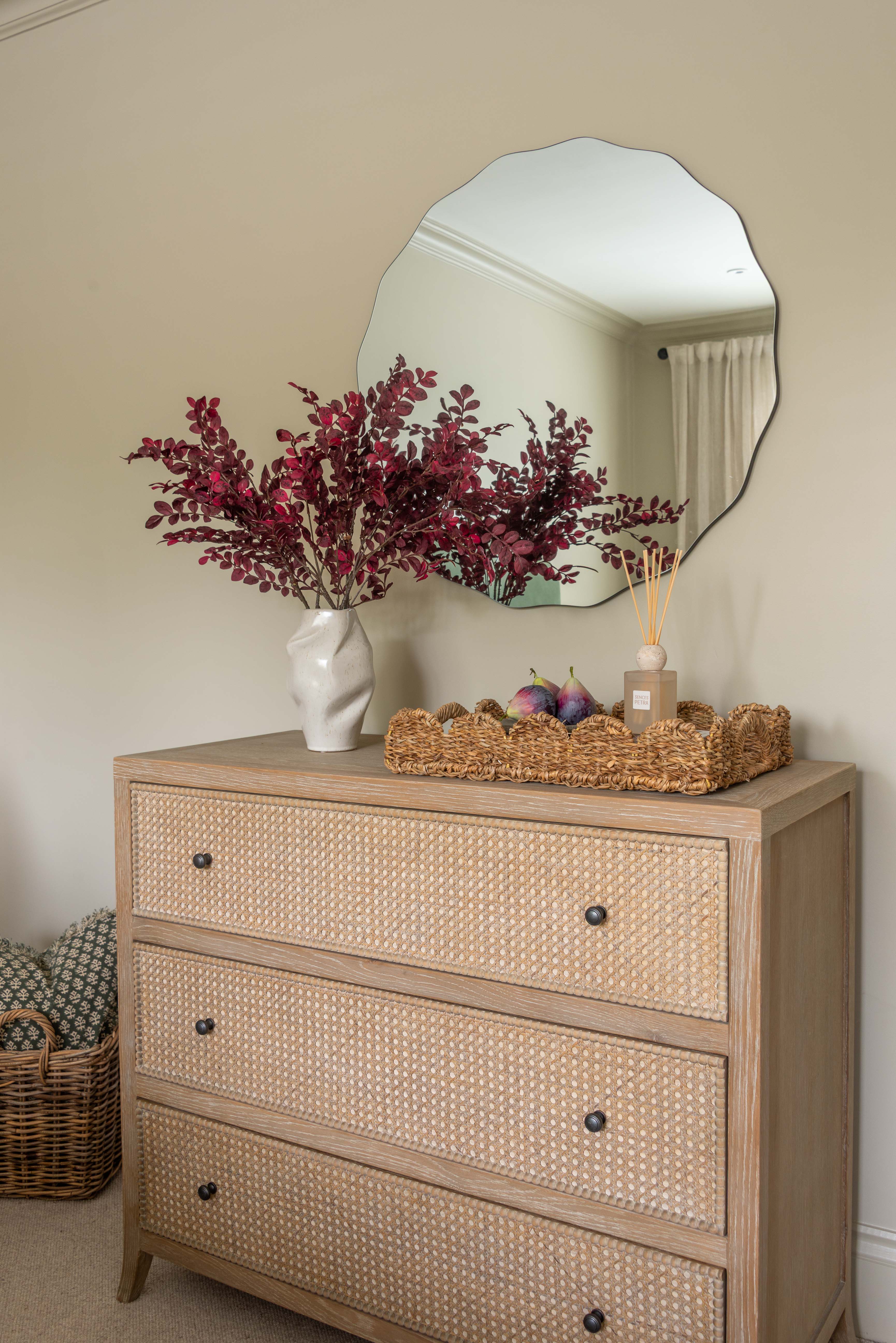 Beige dresser with wicker drawers, decorative flowers, and a mirror on a beige wall.