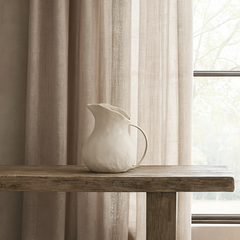 Beige curtain with a wooden shelf and white pitcher in front of a large window.