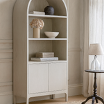 Beige corner bookshelf with decorative items in a room with neutral walls and a lamp.