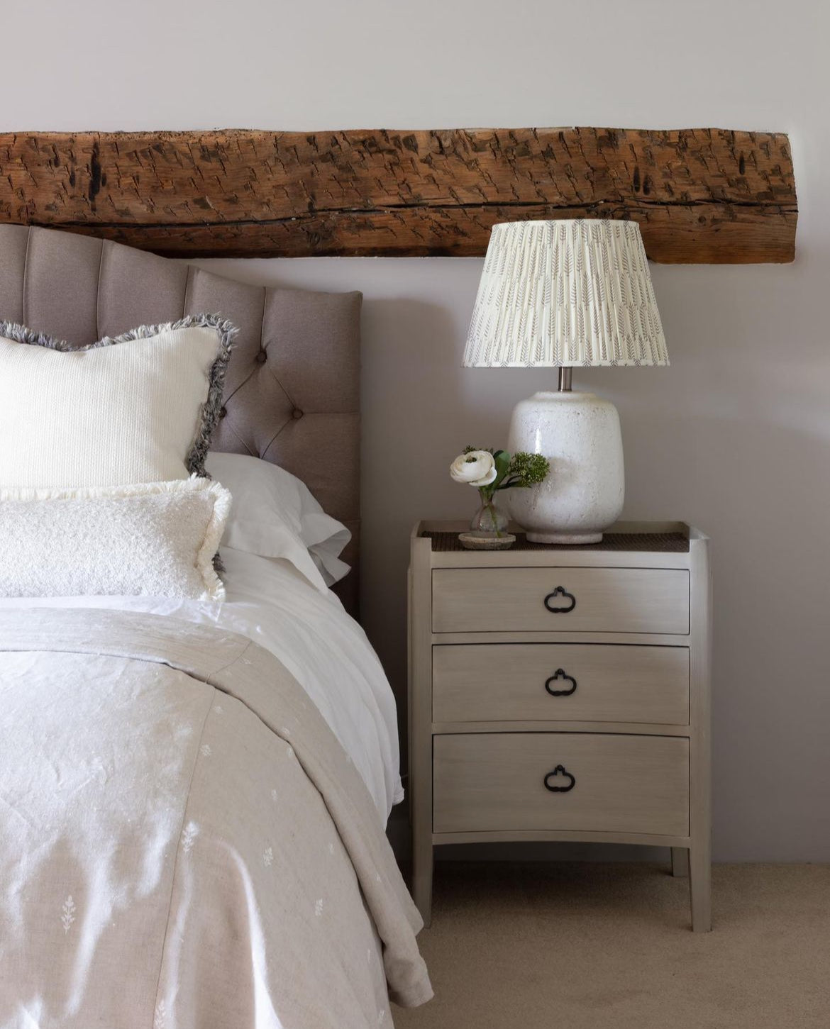 A white wash and rattan bedside table with three drawers, placed next to a bed with a lamp on top.