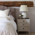 A white wash and rattan bedside table with three drawers, placed next to a bed with a lamp on top.