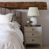 A white wash and rattan bedside table with three drawers, placed next to a bed with a lamp on top.