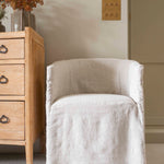 A beige linen dining chair with a low back, seated comfortably in front of a wooden cabinet with flowers on top.