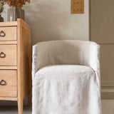 A beige linen dining chair with a low back, seated comfortably in front of a wooden cabinet with flowers on top.