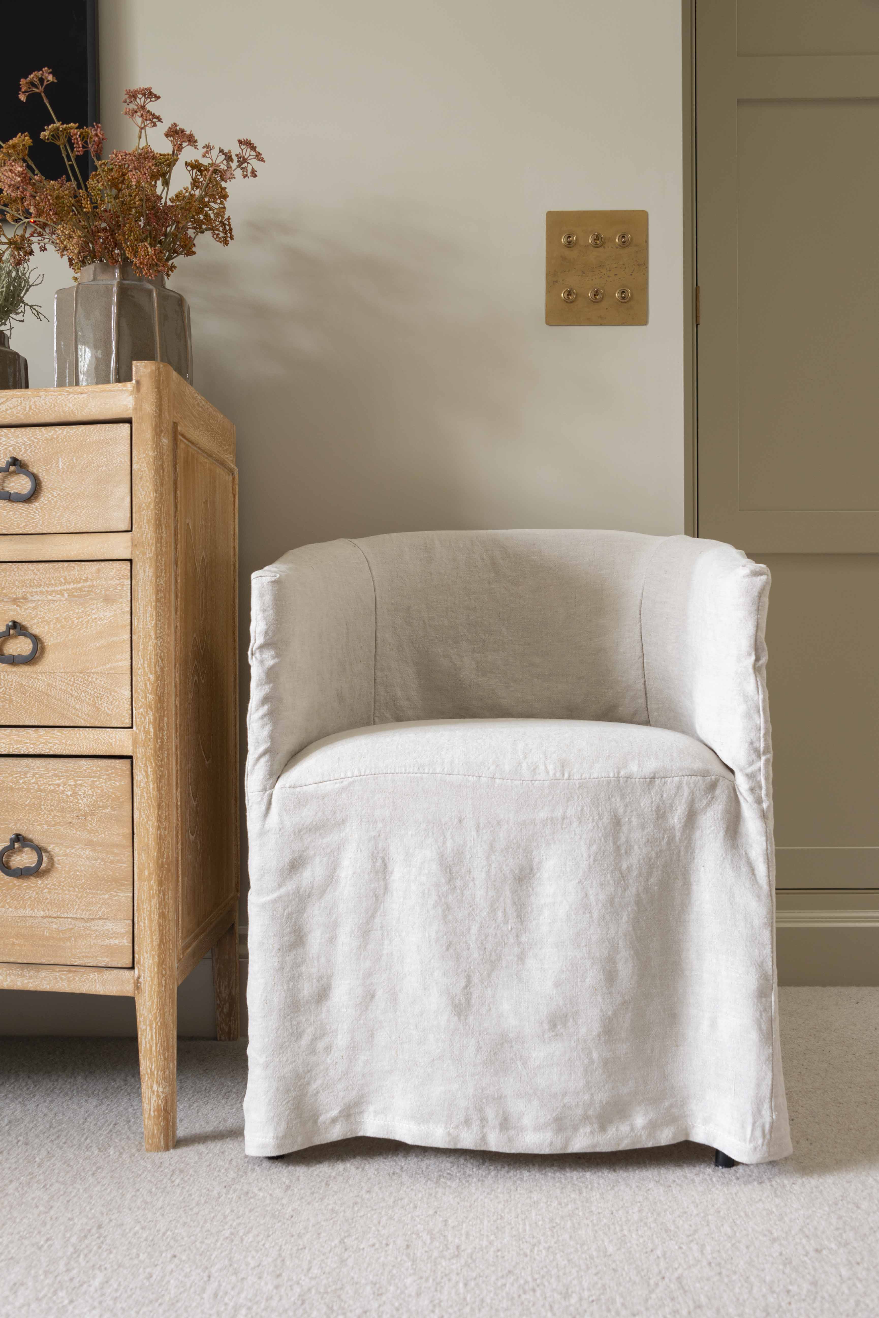 A beige linen dining chair with a low back, seated comfortably in front of a wooden cabinet with flowers on top.