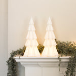 Pair of white lit decorative Christmas trees glowing on a mantelpiece with greenery