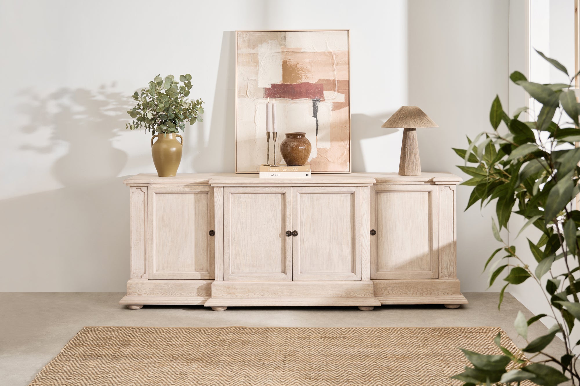 A white oak sideboard with four doors and bronze hardware, placed in a room with decorative items on top and a plant to the side.