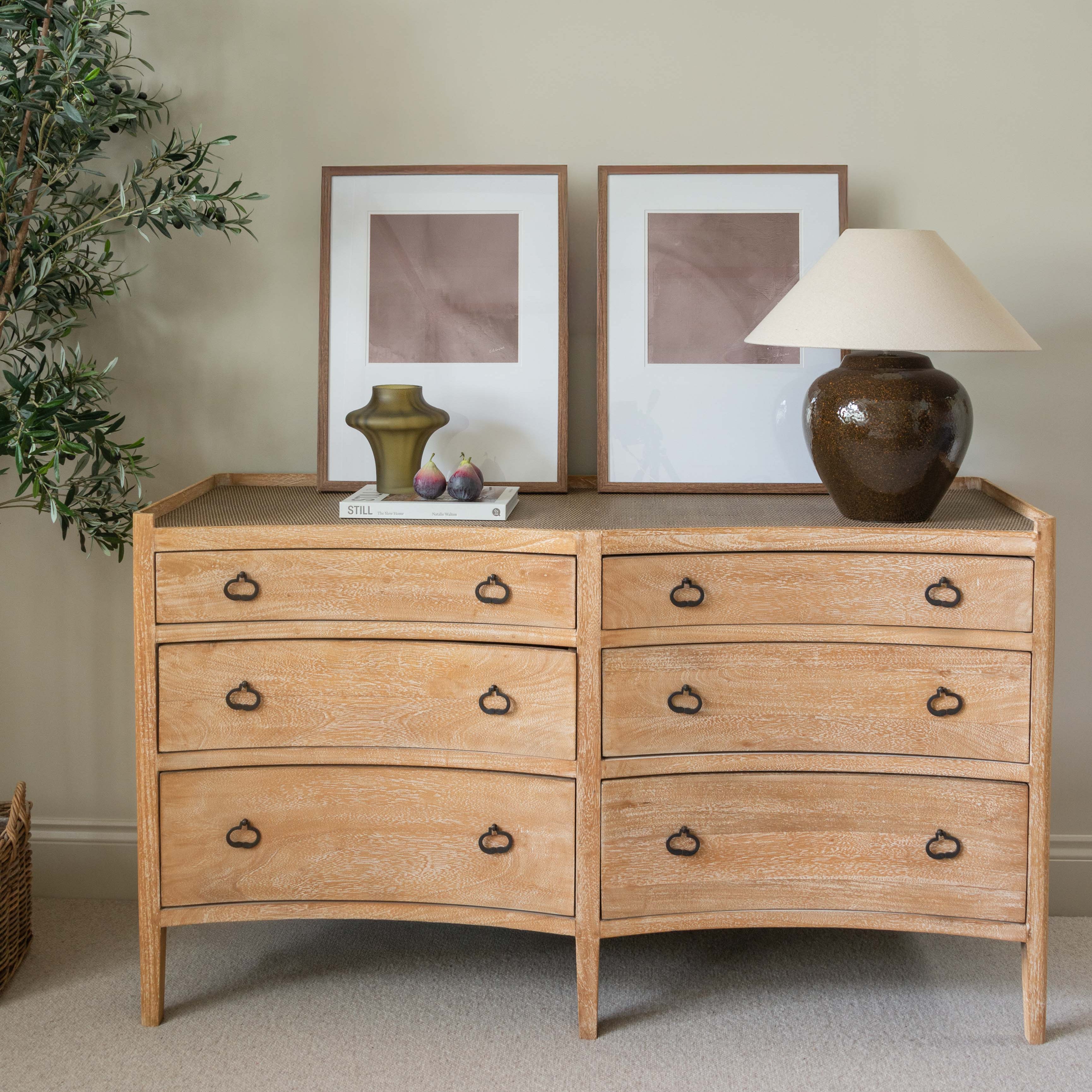 Wooden dresser with two framed pictures and a lamp on top against a beige wall.
