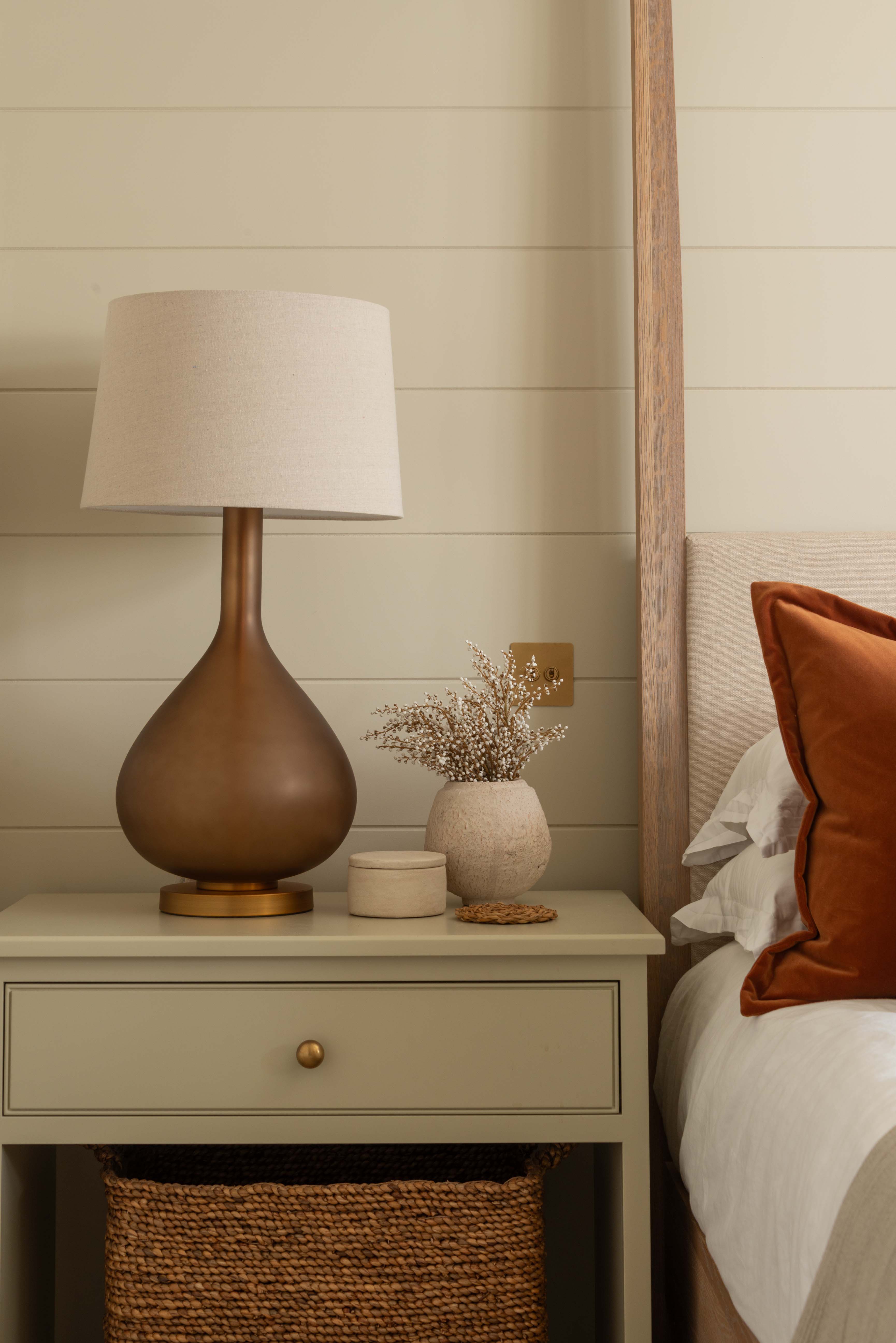 Nighstand with lamp and decorative items next to a bed with orange pillow.