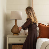 Woman in a brown dress adjusting a lamp on a nightstand in a bedroom.