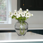 A faux floral arrangement of white ranunculus in a clear glass vase, placed on a kitchen countertop.