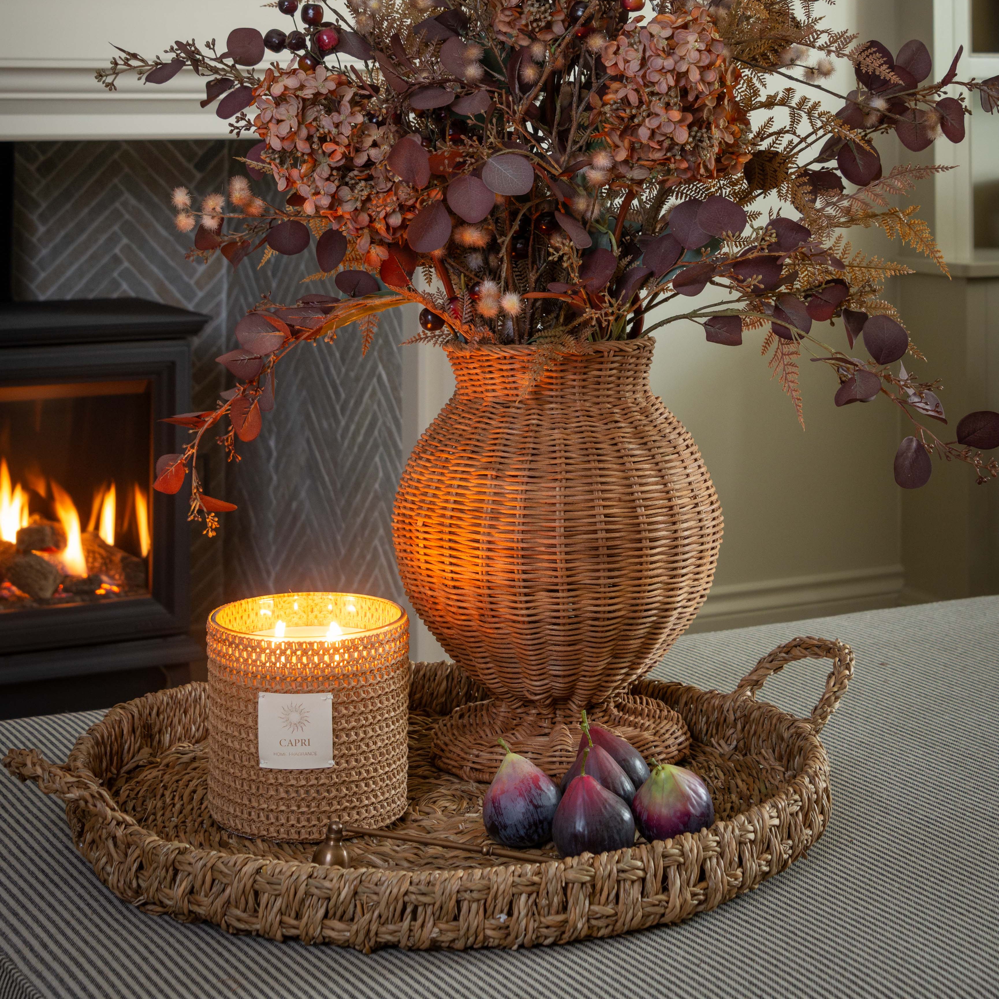 Decorative arrangement with a wicker vase, candle, and figs on a tray in a cozy room.