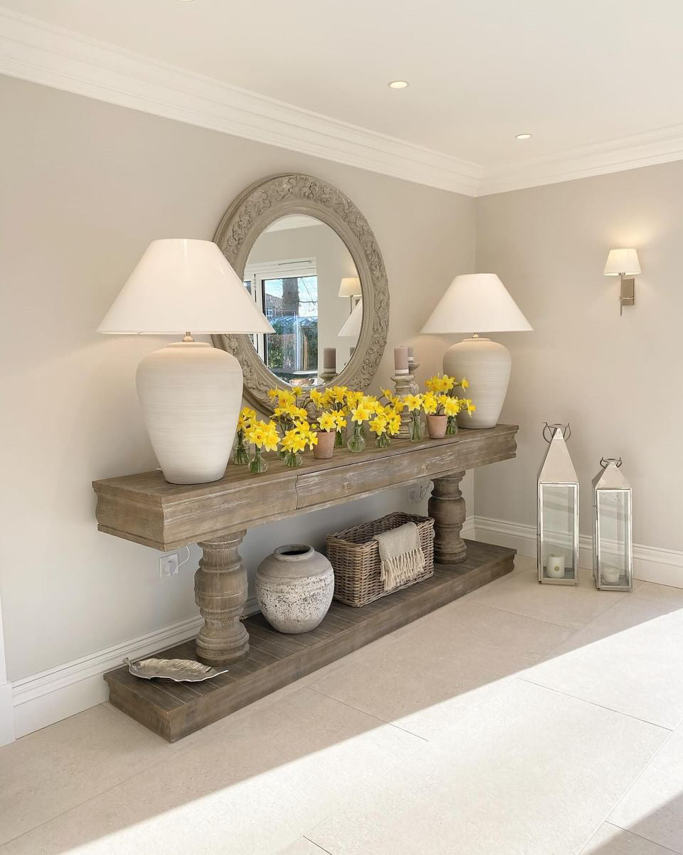 An oversized round mirror with scroll leaf detailing, carved from Paulownia wood, displayed in a room setting with a console table, lamps, and decorative items.
