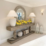 An oversized round mirror with scroll leaf detailing, carved from Paulownia wood, displayed in a room setting with a console table, lamps, and decorative items.