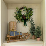 Decorative setup with a wreath, gift boxes, and small trees inside a white cabinet.