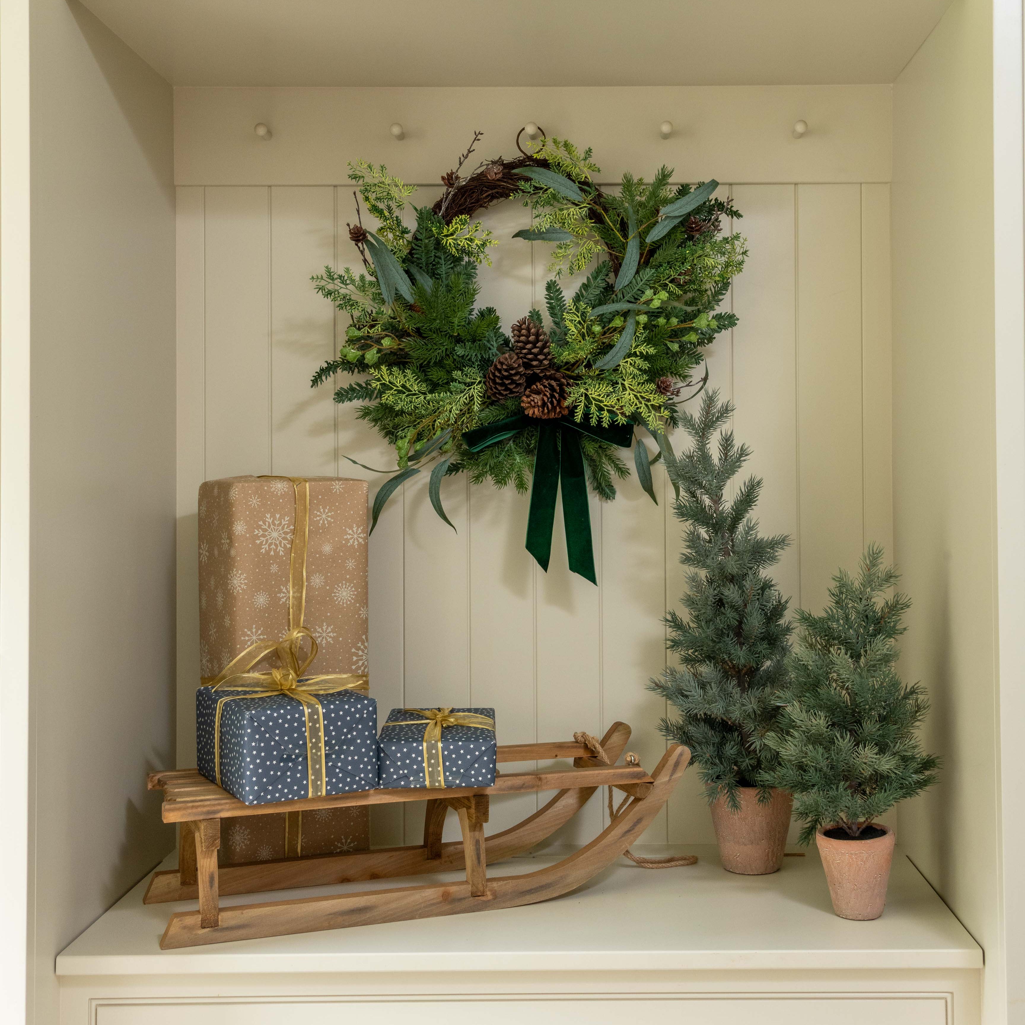 Decorative setup with a wreath, gift boxes, and small trees inside a white cabinet.