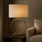 An elegant table lamp with a cream-colored metal ring base and a beige oval lampshade, placed on a side table next to a grey sofa in a dimly lit room.