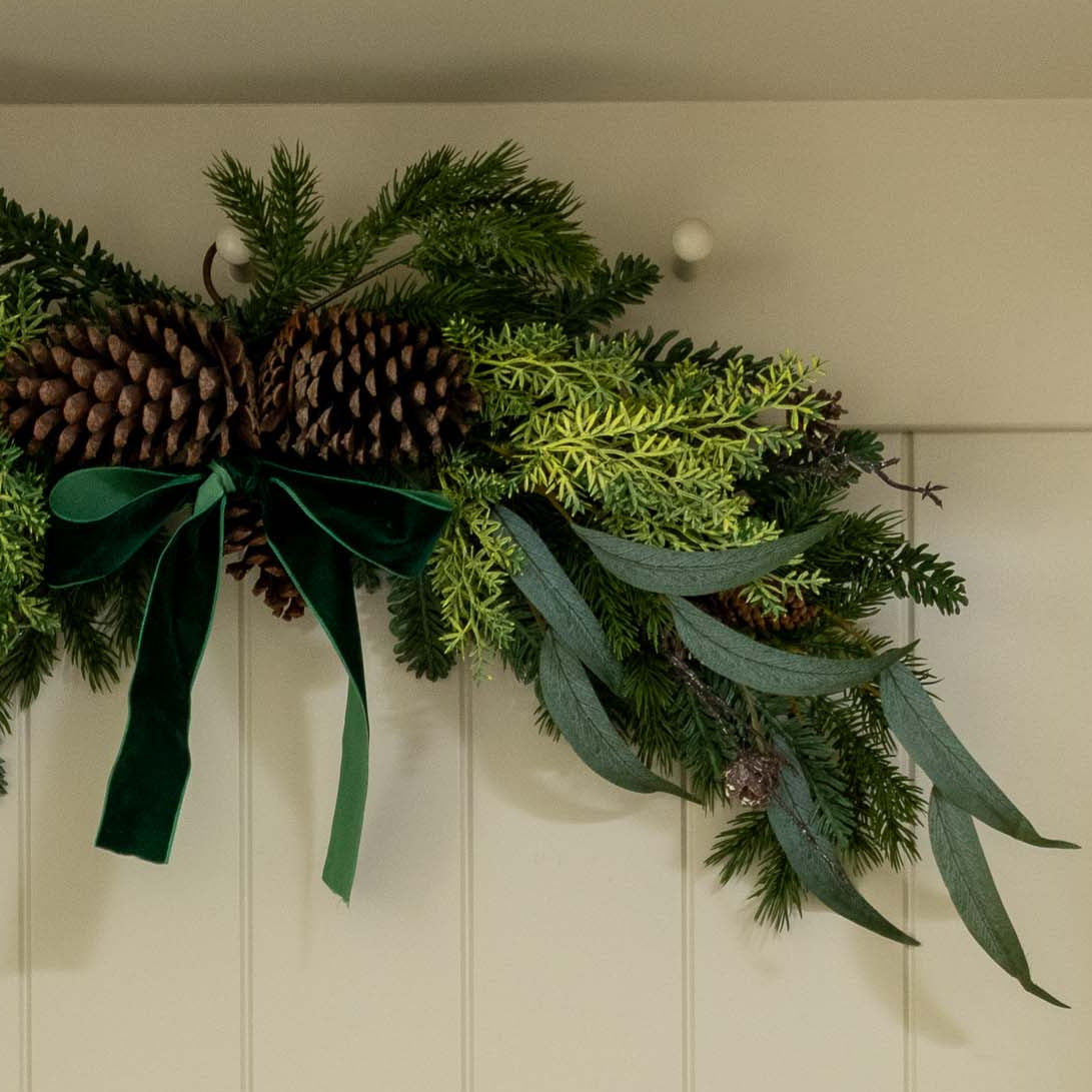 Decorative greenery with pine cones and a green ribbon on a white wall.