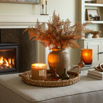 Decorative setting with a vase of dried ferns, candles, and a fireplace in a living room.