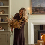 Woman in a long brown dress holding dried fern stems in a cosy living room with candles and a fireplace.