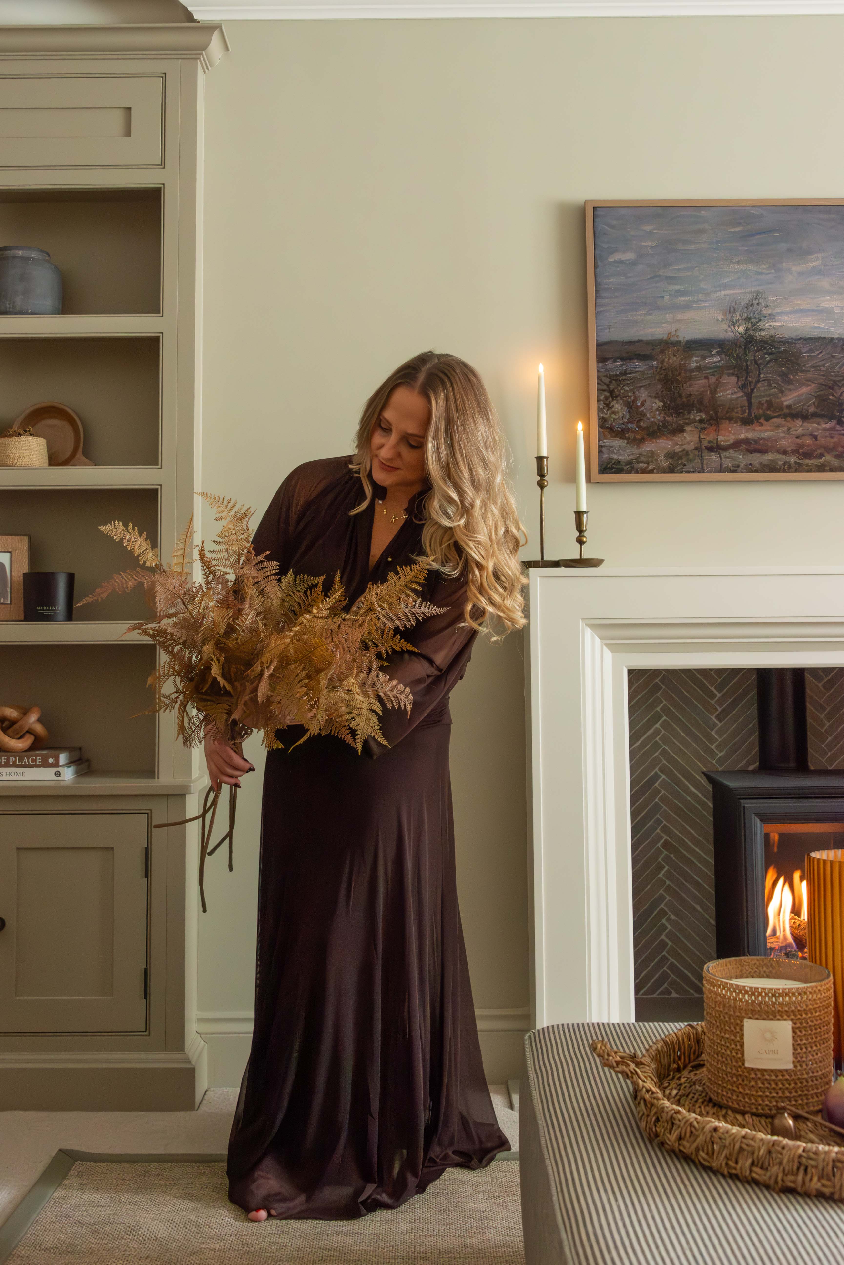 Woman in a long brown dress holding dried fern stems in a cosy living room with candles and a fireplace.