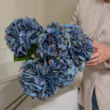 A person holding a faux blue hydrangea stem with realistic petals and leaves.